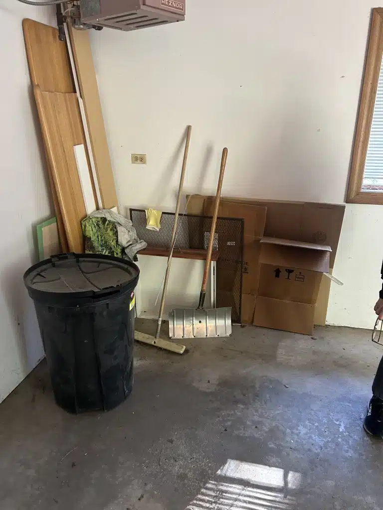 Garage cleanout in Park Ridge, IL 60068, featuring a black plastic trash can, metal shovel, stacked cardboard boxes, and wood in the corner, ready for 24-7JUNK.COM team to clear and dispose of the clutter.